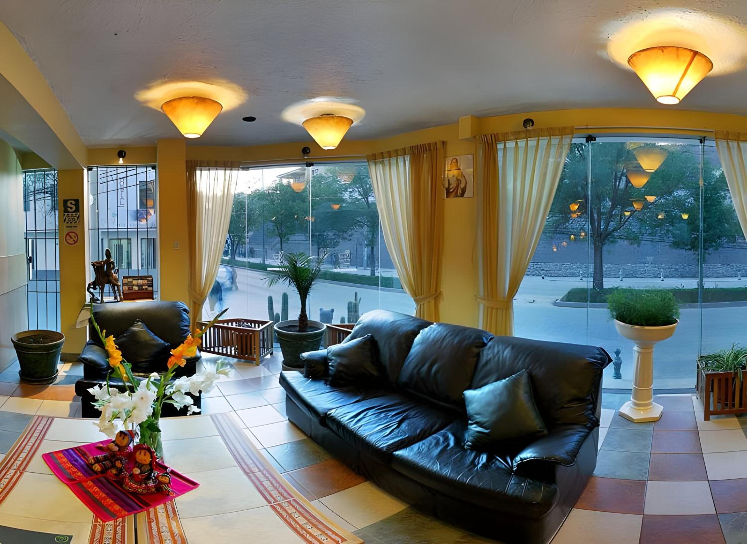 Um hall de hotel moderno com um sofá de couro preto, uma cadeira de madeira e uma mesa pequena com um vaso de flores. Grandes janelas com cortinas oferecem vista para a rua.