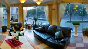 A modern hotel lobby with a black leather sofa, a wooden chair, and a small table with a vase of flowers. Large windows with curtains provide a view of the street outside. - El Puma (Cusco)
