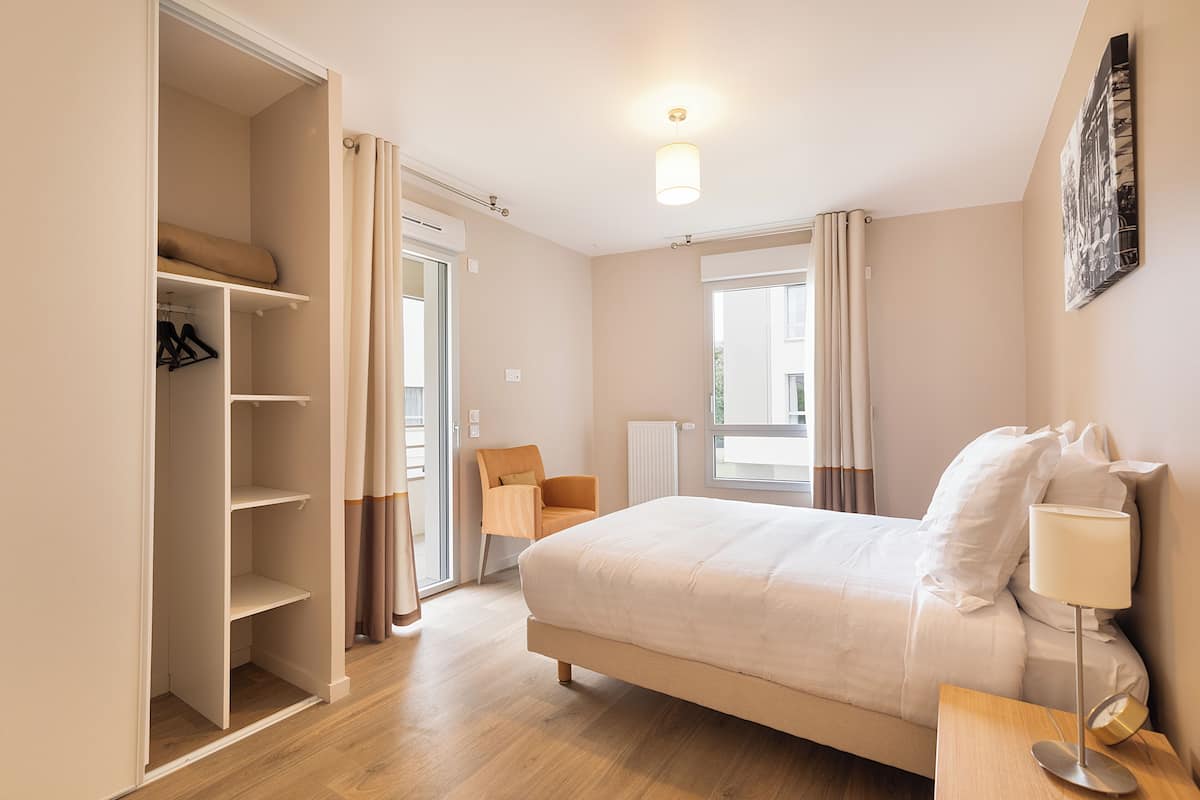A bedroom with a bed, a chair, a bedside table, a lamp, a wardrobe, and a window with curtains.