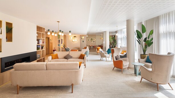 A spacious living room with a sofa, armchairs, and a TV. There are bookshelves and a large plant. - Zenao Appart'hôtels Montreuil (Montreuil)