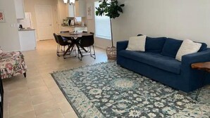 Een woonkamer met een blauwe bank, een vloerkleed met patroon en een eethoek met tafel en stoelen.