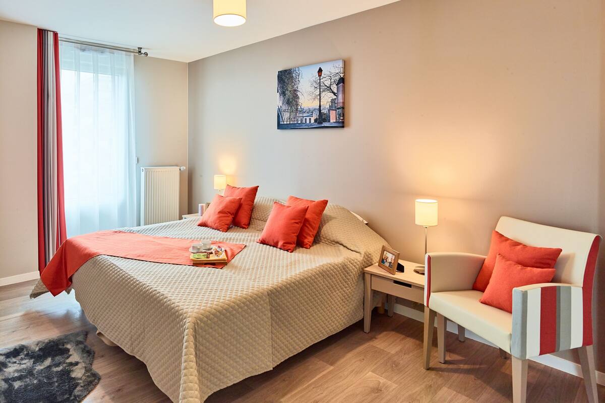 Quarto com cama, mesas de cabeceira, uma cadeira, radiador, janela com cortinas e um quadro na parede.
