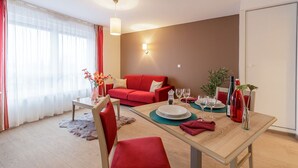 Una sala de estar moderna con un sofá rojo, una mesa de comedor para dos personas y un ventanal con cortinas traslúcidas.