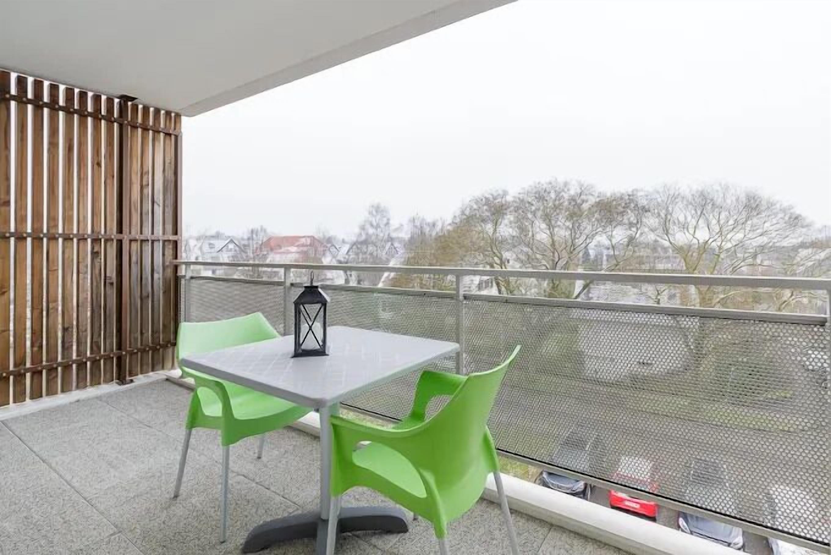 A balcony with a table and chairs, a lantern, and a view of trees and buildings.