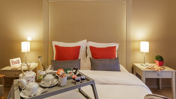 Habitación de hotel con cama, mesitas de noche, una mesita con alimentos para el desayuno y una mesita de noche con lámpara y planta.