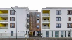Un edificio moderno de varios pisos con balcones, una entrada central y un pequeño árbol en la parte delantera.