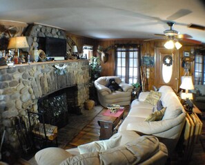 Un salon chaleureux avec une cheminée en pierre, un canapé, un fauteuil et un ventilateur de plafond.