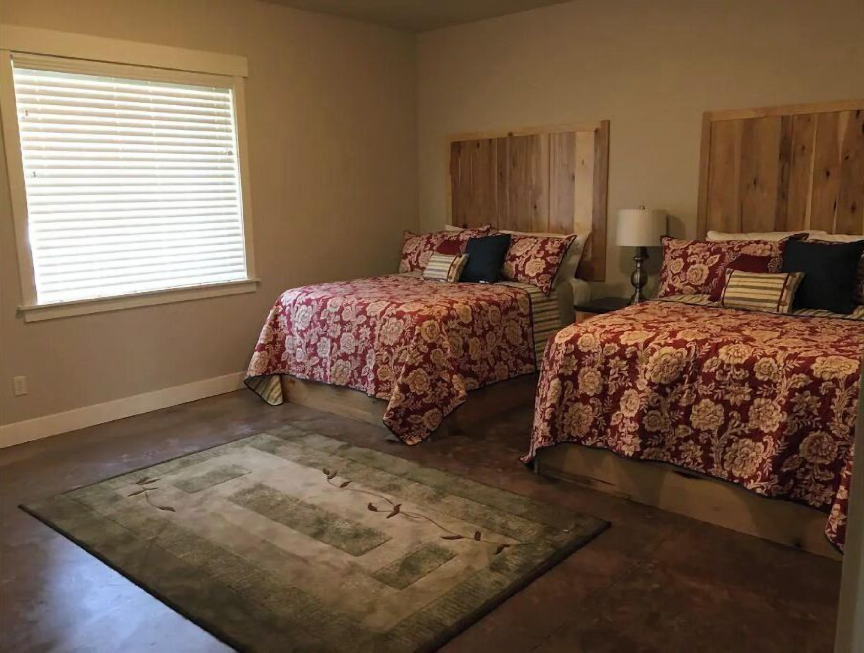A bedroom with two beds, a large window with blinds, a wooden headboard, a nightstand with a lamp, and a green area rug.
