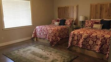 A bedroom with two beds, a large window with blinds, a wooden headboard, a nightstand with a lamp, and a green area rug.