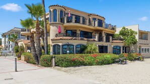 A multi-story building with balconies, palm trees, and a sandy area with benches.