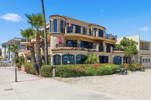 A multi-story building with balconies, palm trees, and a sandy area with benches.