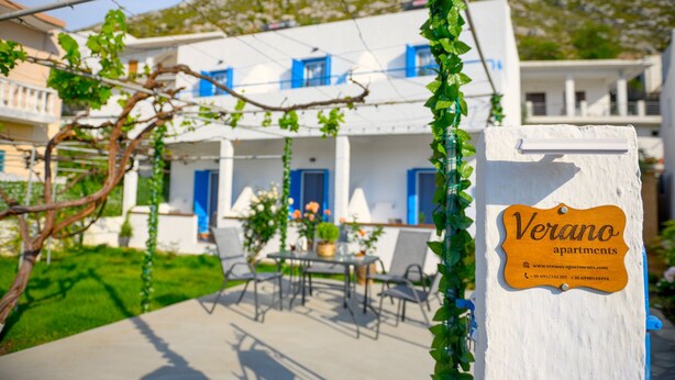 Een wit gebouw met blauwe luiken, een bordje met de tekst 'Verano apartments' en een tuin met stoelen en tafels.