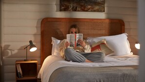 Una persona llegint un llibre al llit, amb coixinets blancs, un capçal de fusta i una tauleta de nit amb una làmpada i llibres.