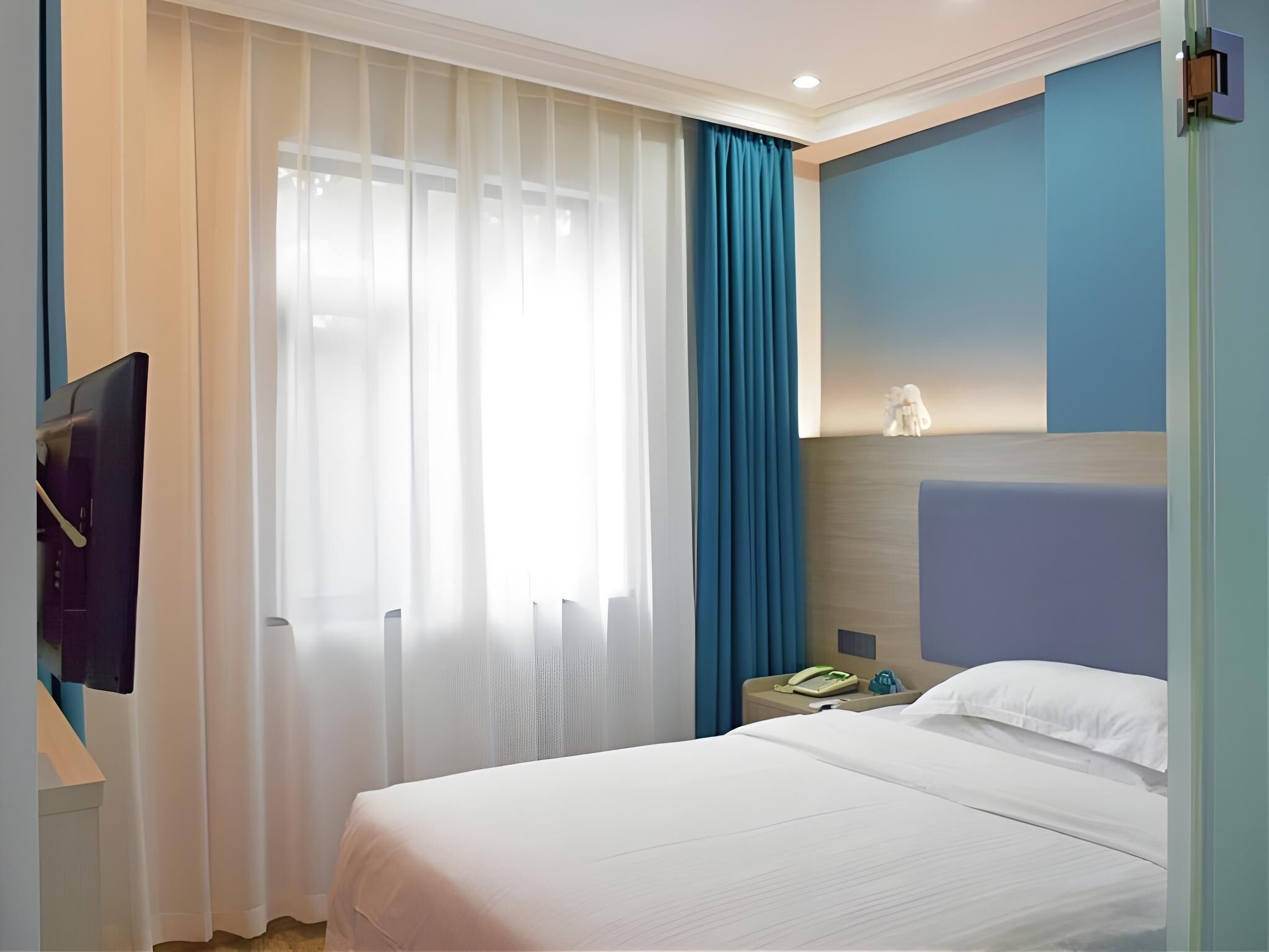 A hotel room with a bed, a TV mounted on the wall, a nightstand with a phone, and a window with sheer curtains and a blue drape.