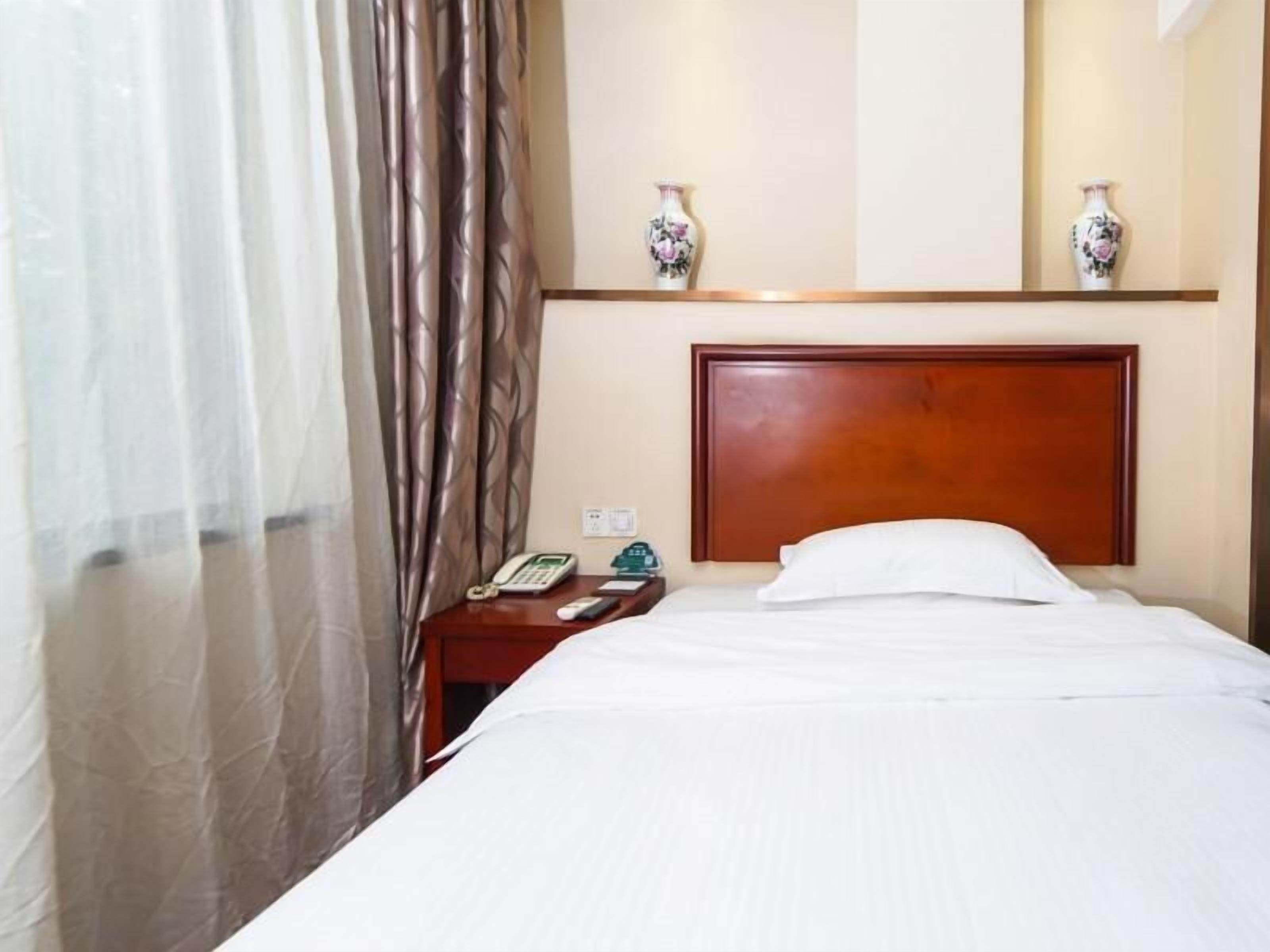 A hotel room with a bed, a wooden headboard, a nightstand with a phone and a remote control, and two decorative vases on a shelf above the bed.
