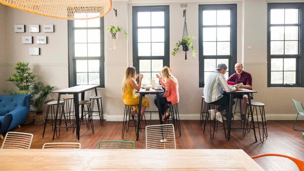 Un coin repas avec une table en bois, des tabourets de bar et un luminaire suspendu. On y trouve également des plantes en pot et un canapé bleu.