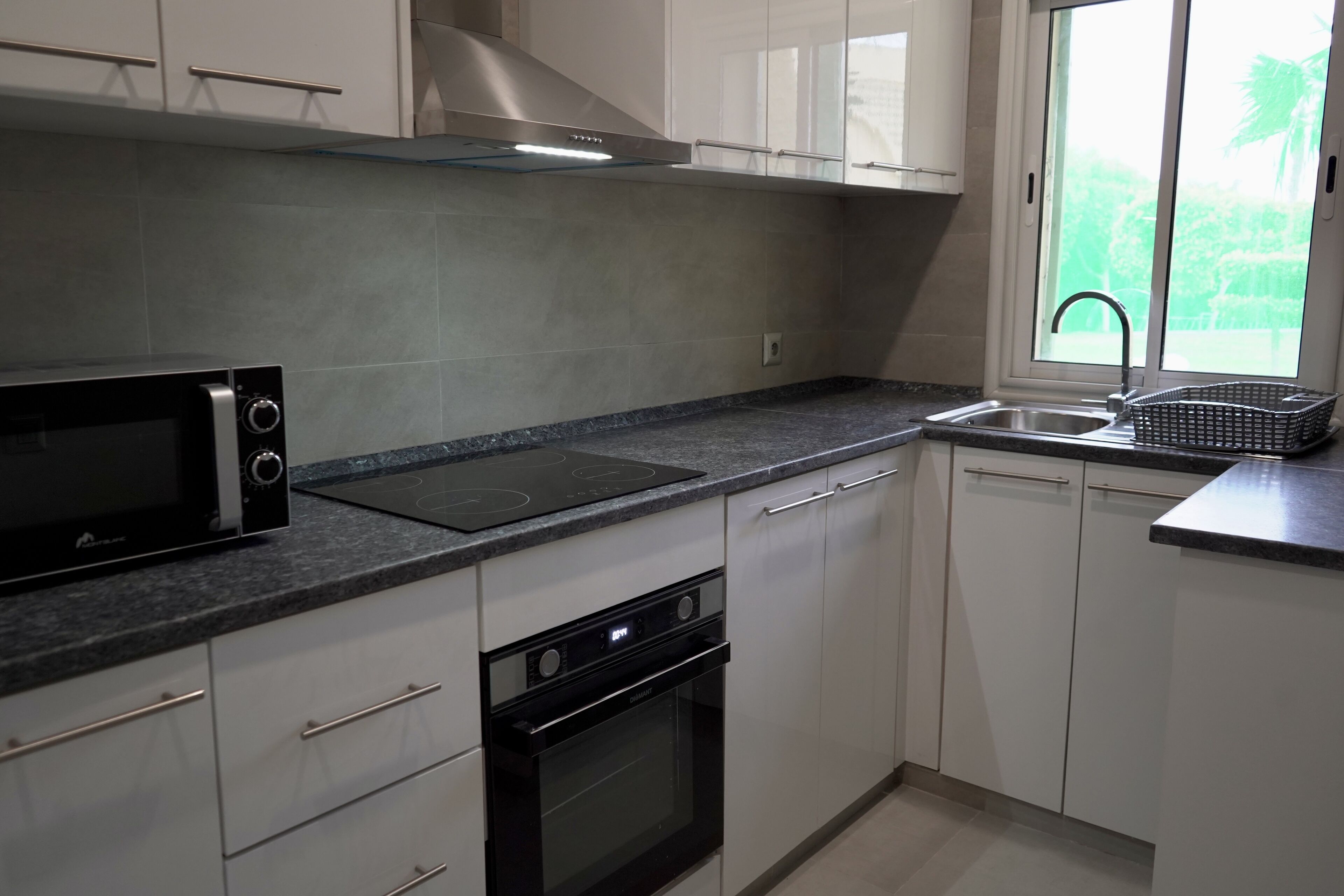 Een moderne keuken met witte kasten, een zwarte magnetron, een oven, een kookplaat en een raam met uitzicht op groen.