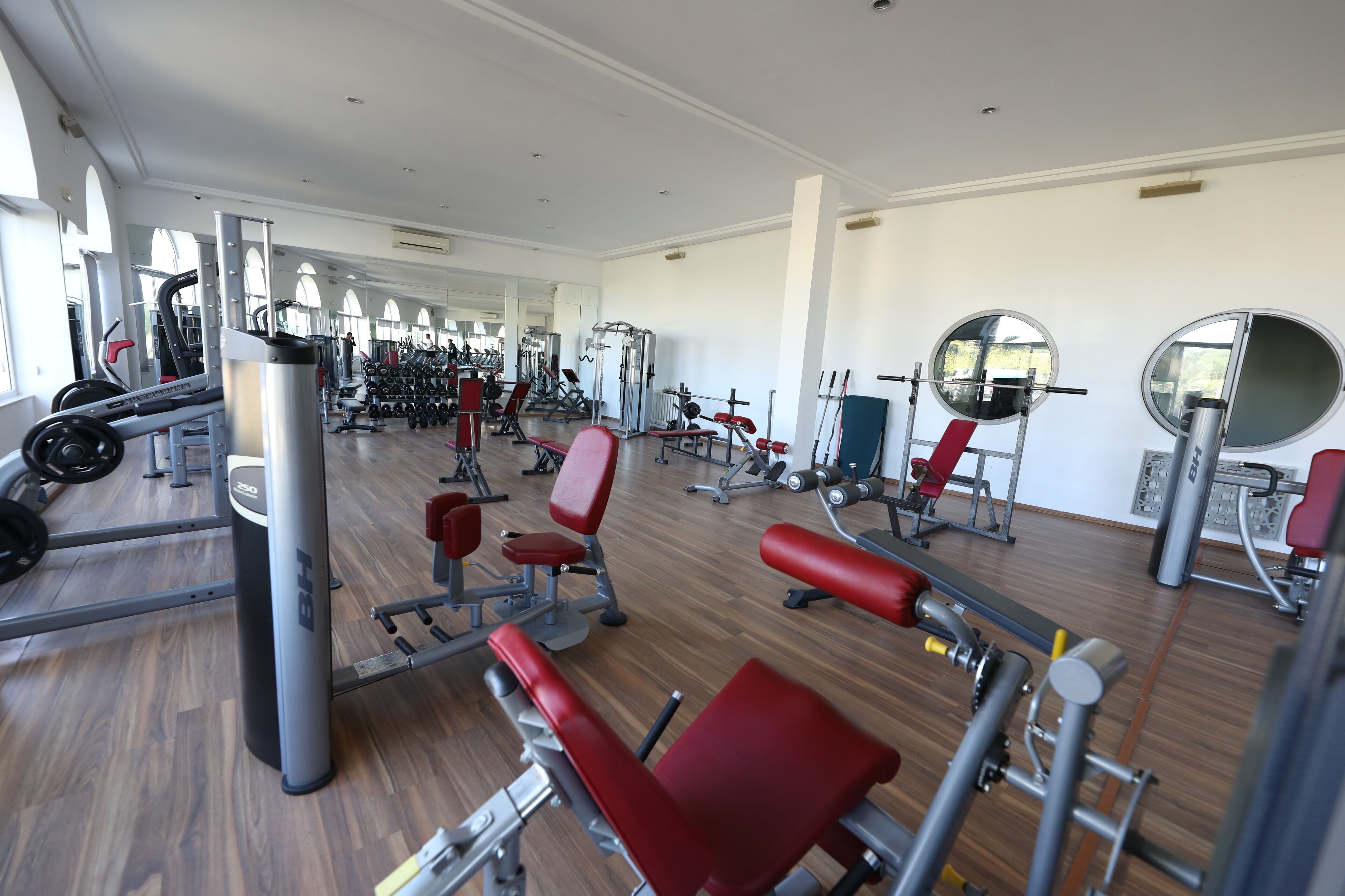A gym with various exercise equipment, including weight machines and free weights, arranged in a spacious room with large windows.