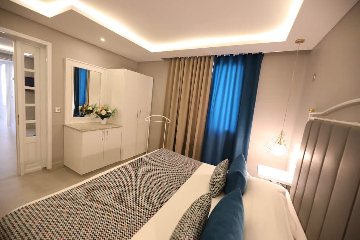 A modern bedroom with a large bed, a wardrobe, a vanity with a mirror, and a window with curtains.