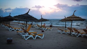 Een strand met rijen ligbedden en rieten parasols, een reddingsboei en een rustige zee onder een zonsondergang.