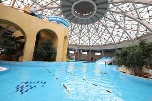 Ein Hallenbad mit deutlich markierten Bahnen, einer zentralen Rutsche und einer hohen, geometrischen Glasdecke.