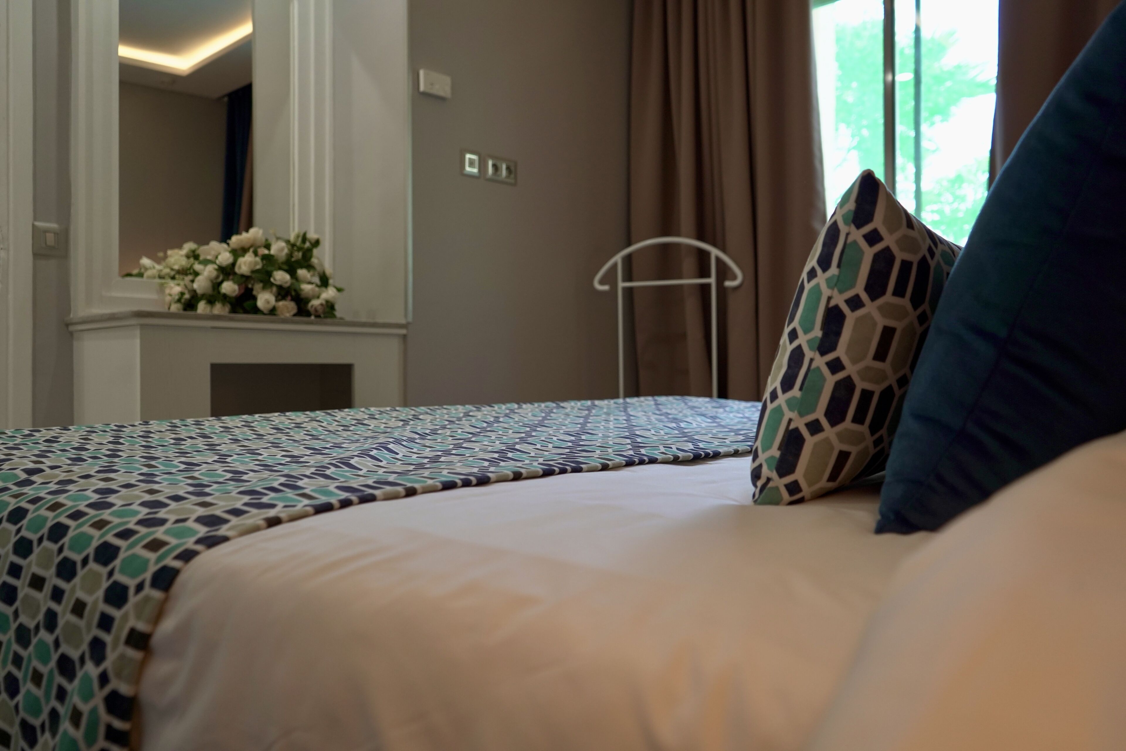 Una cama bien tendida con ropa de cama estampada, una mesita de noche y un espejo que refleja una habitación con chimenea y una ventana con cortinas.