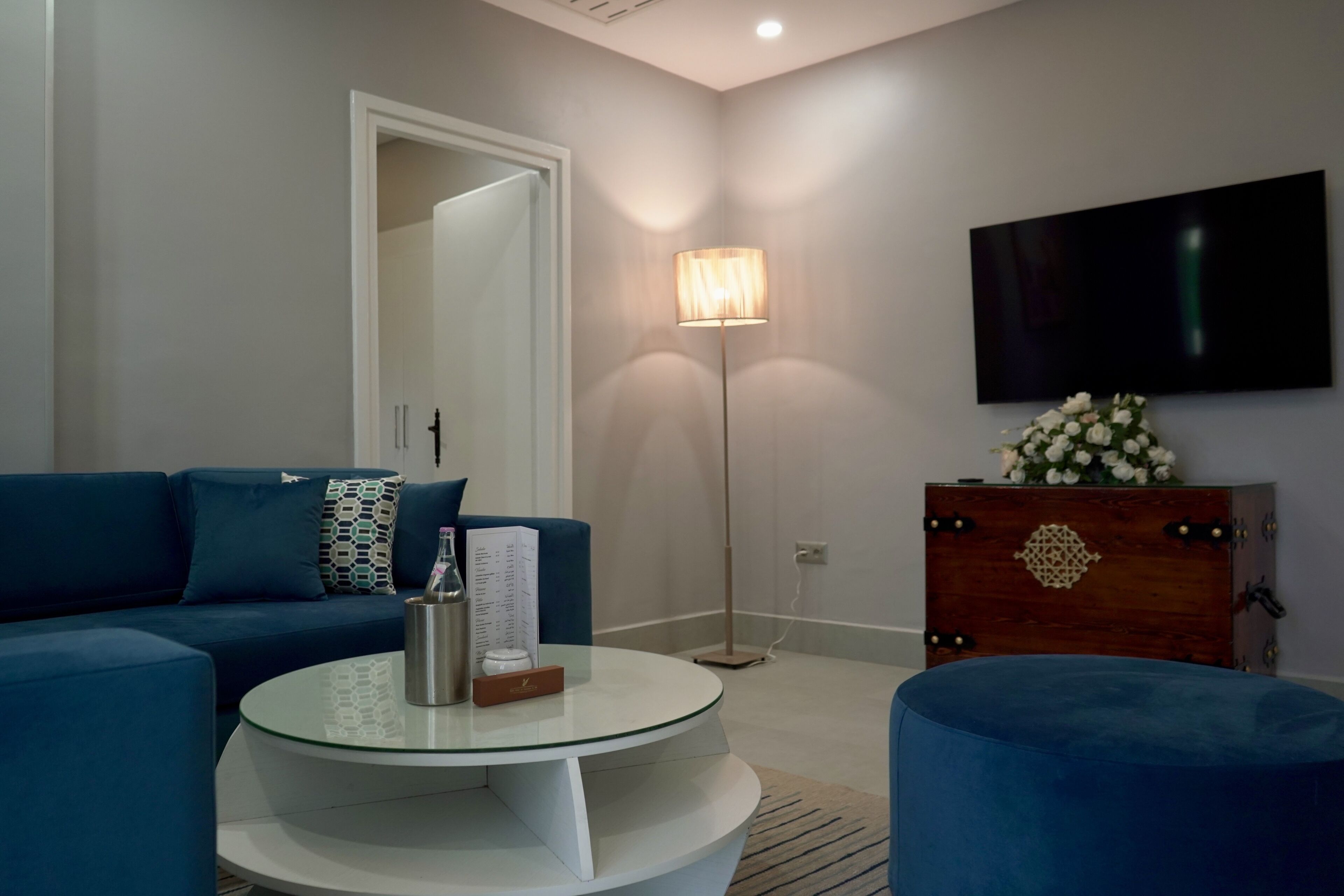 Una sala moderna con un sofá azul, una mesa de centro de vidrio, un aparador de madera con un arreglo floral y una lámpara de pie.