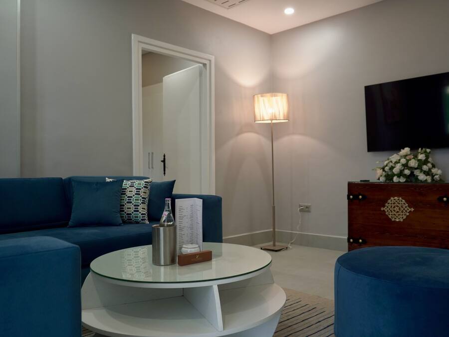 Una sala moderna con un sofá azul, una mesa de centro de vidrio, un aparador de madera con un arreglo floral y una lámpara de pie.