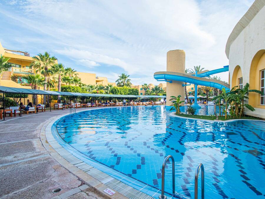 En stor inomhuspool med en böjd design, omgiven av palmer och ett avskärmat sittområde.