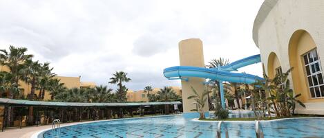 A swimming pool with a water slide, palm trees, and a covered area.