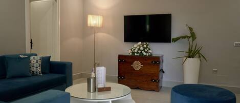 A modern living room with a blue sofa, a white coffee table, a round white side table, a flat-screen TV, a wooden chest of drawers, and a potted plant.