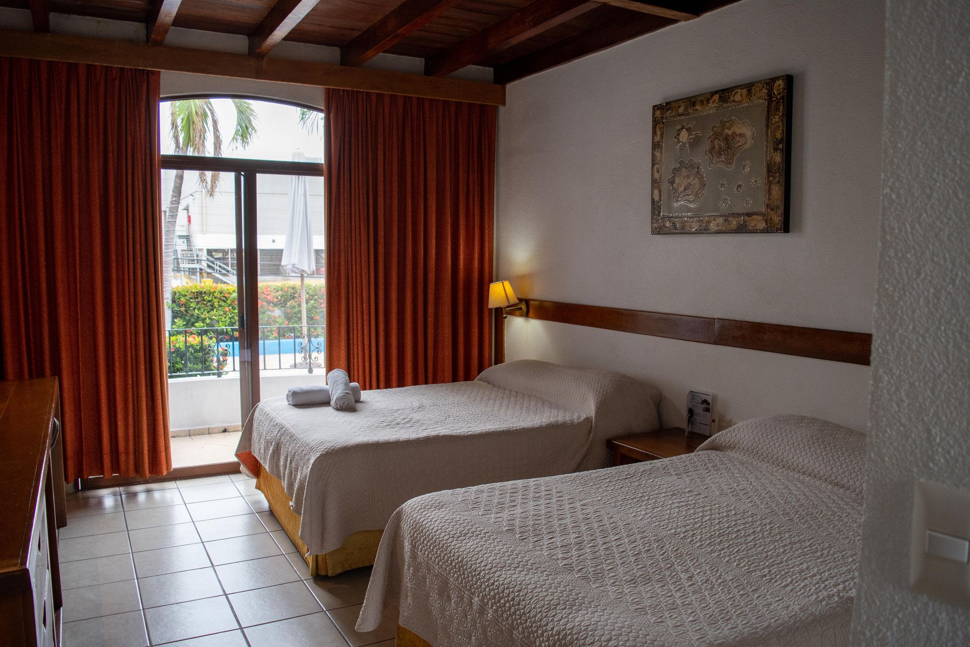 Una habitación de hotel con dos camas, un balcón con cortinas rojas, un cuadro en la pared y una lámpara en la mesita de noche.