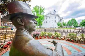 A statue of a man in a hat pointing to a building in the background.