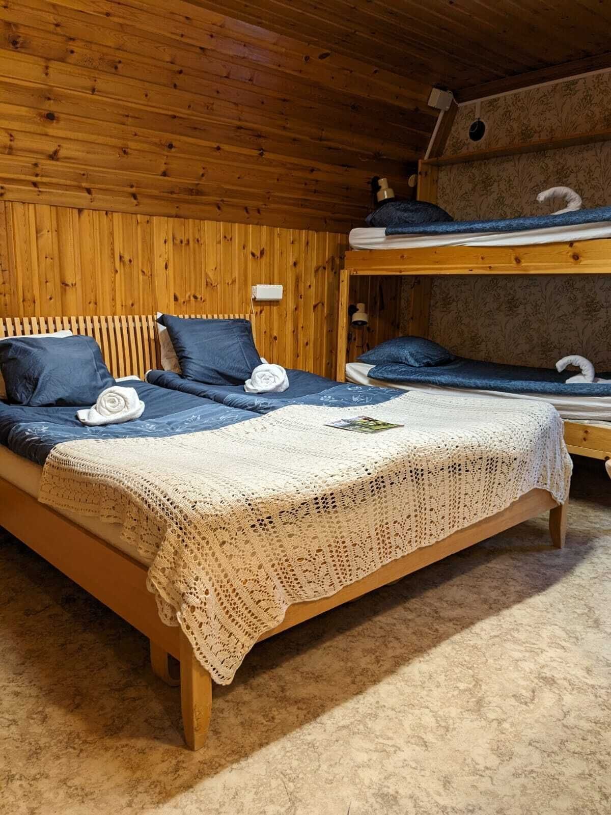 Una camera in legno arredata con un letto a castello, un letto singolo con una coperta bianca lavorata a maglia e un tavolino con una lampada.