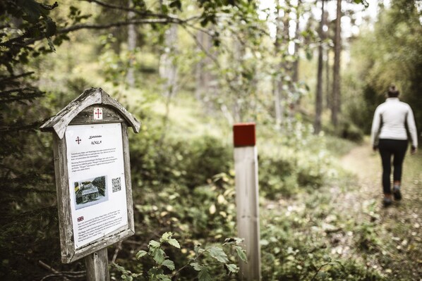 A wooden signpost with a QR code and text, a red post, and a forested path with a person walking away. - Hotel Nederhögen Vildmarkscenter (Rätan)