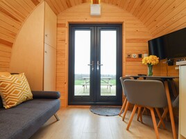 Una cabaña acogedora con interior de madera, un sofá con un cojín estampado, una mesa de comedor con sillas y un ventanal con vistas al exterior.