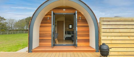 A cozy, semi-open wooden structure with a curved roof, a wooden deck in front, and a barbecue grill to the right.