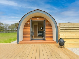 A cozy, semi-open wooden structure with a curved roof, a wooden deck in front, and a barbecue grill to the right.