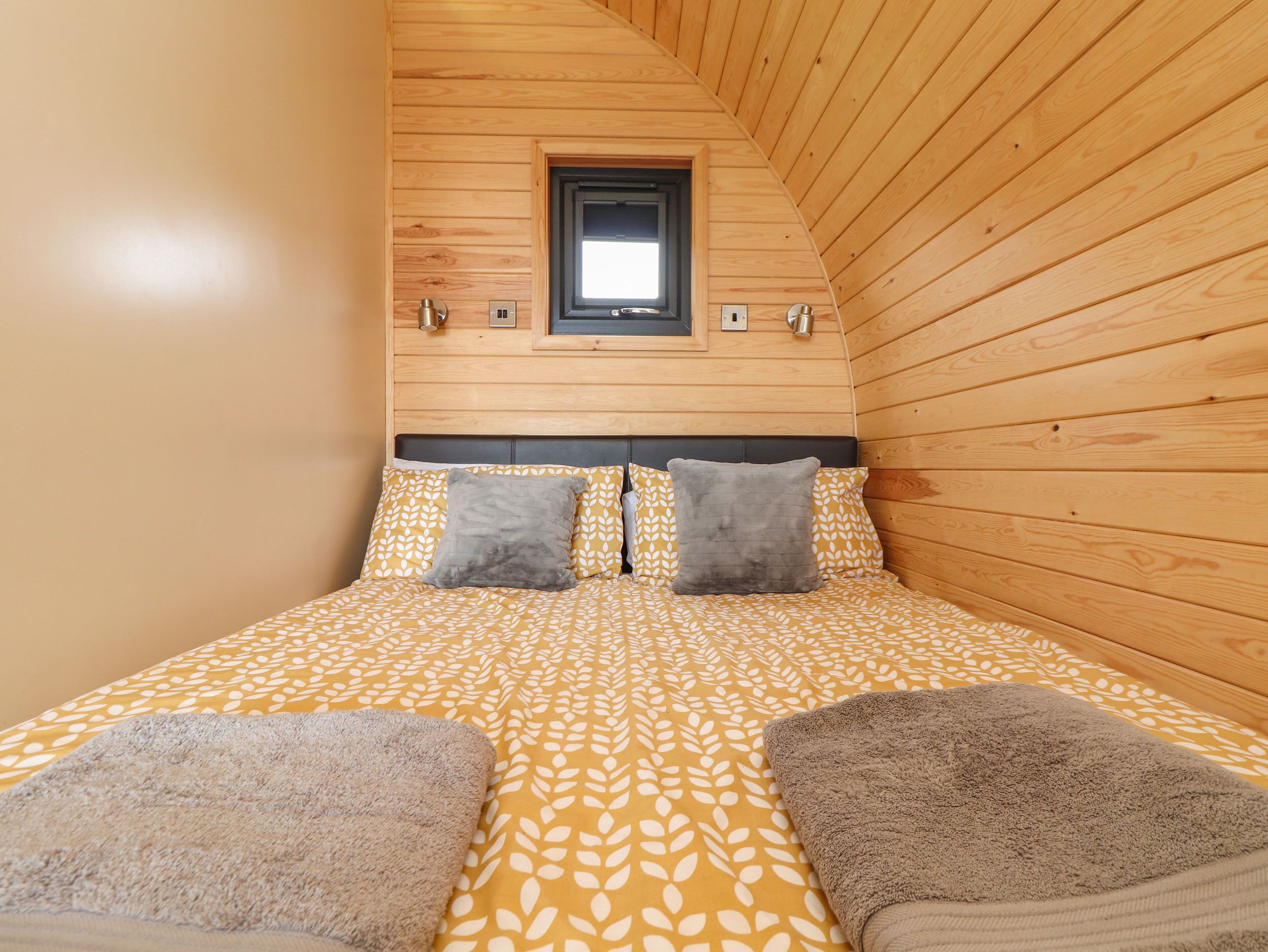 Ein Schlafzimmer mit schrägen Wänden, Holzwänden, einem Bett mit gemusterten Bettbezügen, zwei Kissen und einem kleinen Fenster.