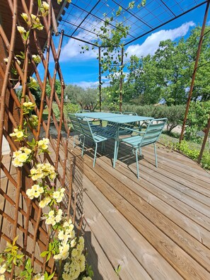 Eine Holzterrasse mit einem Metalltisch und -stühlen, umgeben von viel Grün und einem Metall-Rankgerüst mit Blumen.