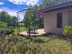Eine Terrassenanlage mit Pergola, Tisch und Stühlen, umgeben von Grünpflanzen und einem Haus.