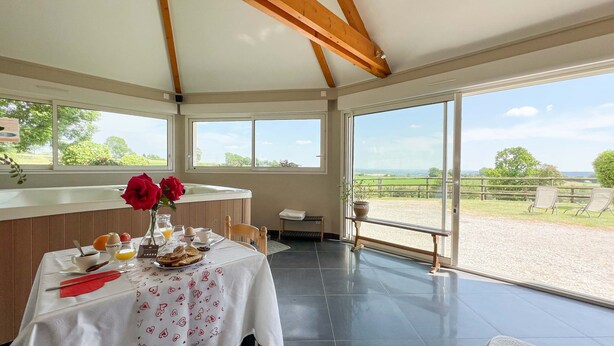 Un comedor con una mesa dispuesta para el desayuno, ventanas amplias que ofrecen vistas al campo, y una estructura de techo de madera.