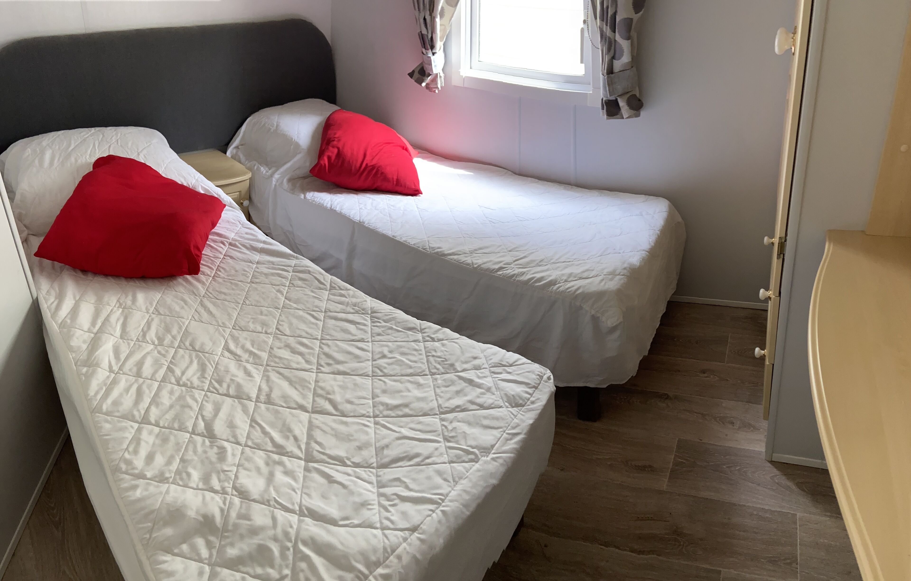 Ein Zimmer mit zwei Einzelbetten, jeweils mit einem roten Kopfkissen, einem Nachttisch und einem Vorhangfenster.