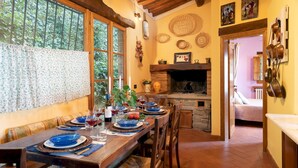A dining area with a long wooden table set for a meal, a fireplace, and a kitchen area with hanging pots. - Podere Belvedere 6, Emma Villas (Montevarchi)