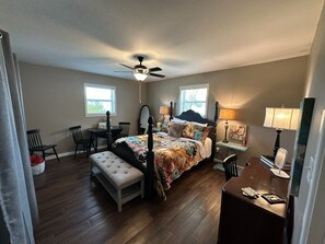 A bedroom with a bed, a desk, a chair, a bench, a ceiling fan, and two windows.