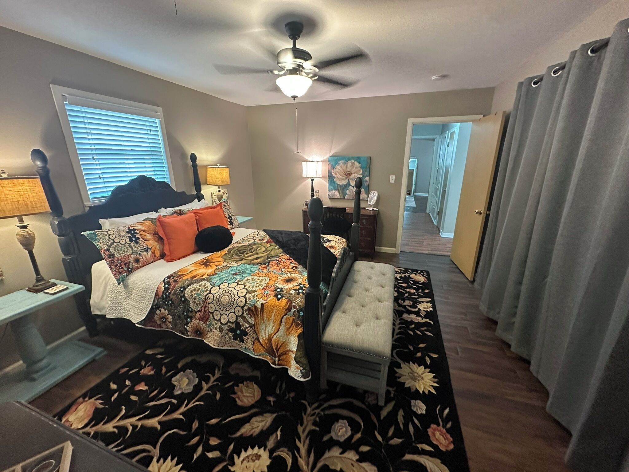 A bedroom with a bed, a nightstand, a lamp, a ceiling fan, a window with blinds, a door, and a hallway.