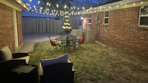 En trädgård med ett bord och stolar på en uteplats, ett randigt parasoll och ljusslingor.