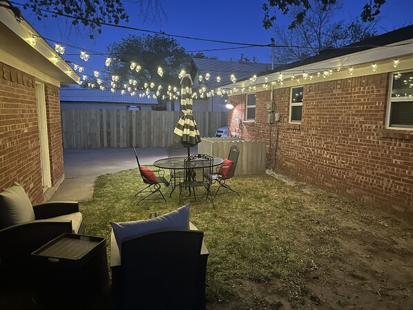 A backyard with a patio table and chairs, a striped umbrella, and string lights.