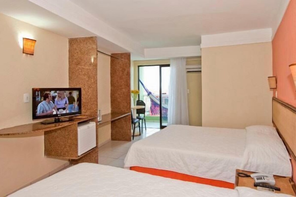 A hotel room with two beds, a television, a desk, and a view of the outside.