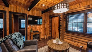 Interior acogedor de una cabaña de troncos con un sofá de cuero, una mesa de centro redonda de madera, una chimenea y un ventanal grande con persianas.